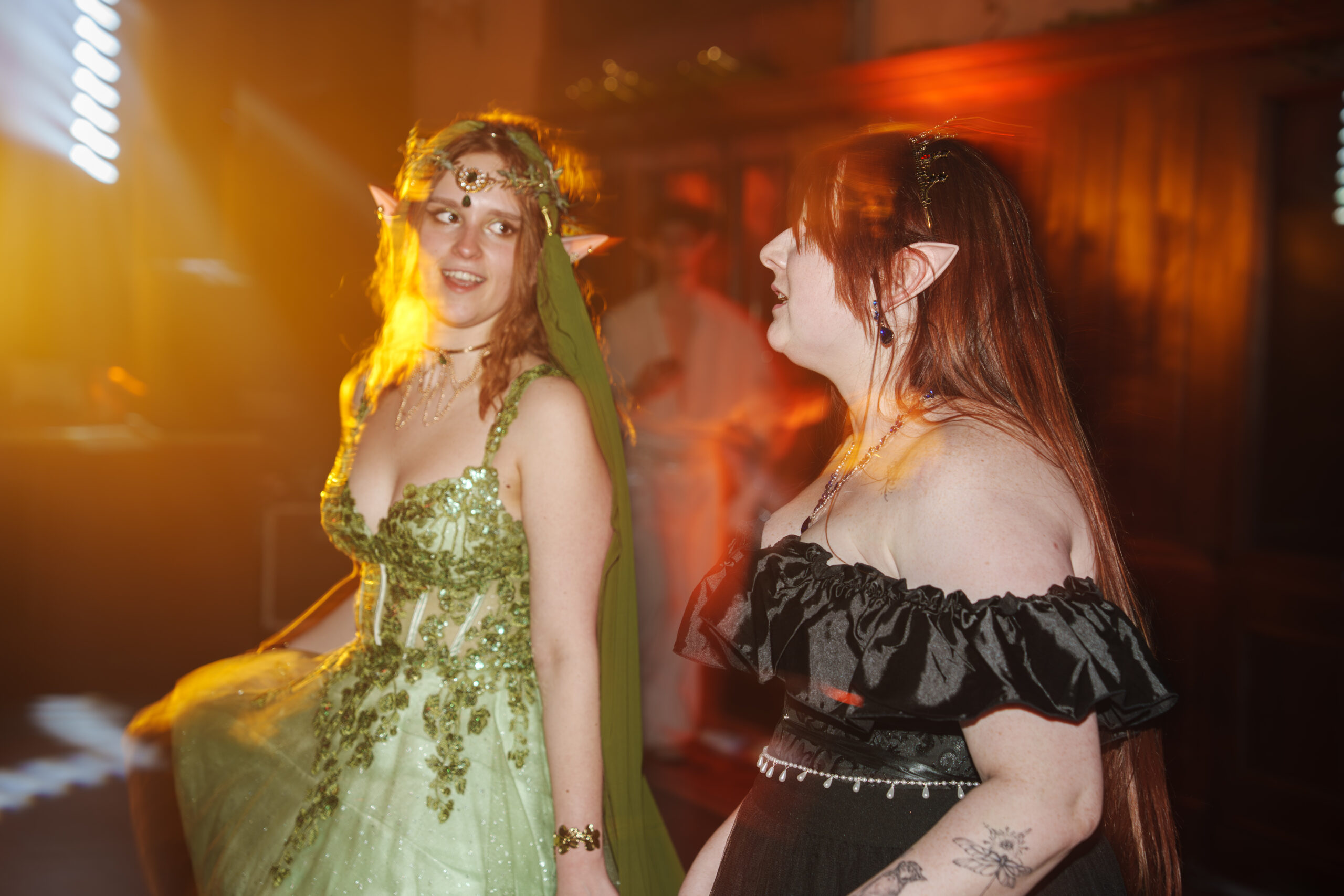 Two women in elf costumes at a glowing, warm-lit party, one in a gold-green beaded gown with a leafy headpiece and pointy ears, the other in a black off-shoulder dress with tattoos visible on her arm, both chatting or singing.