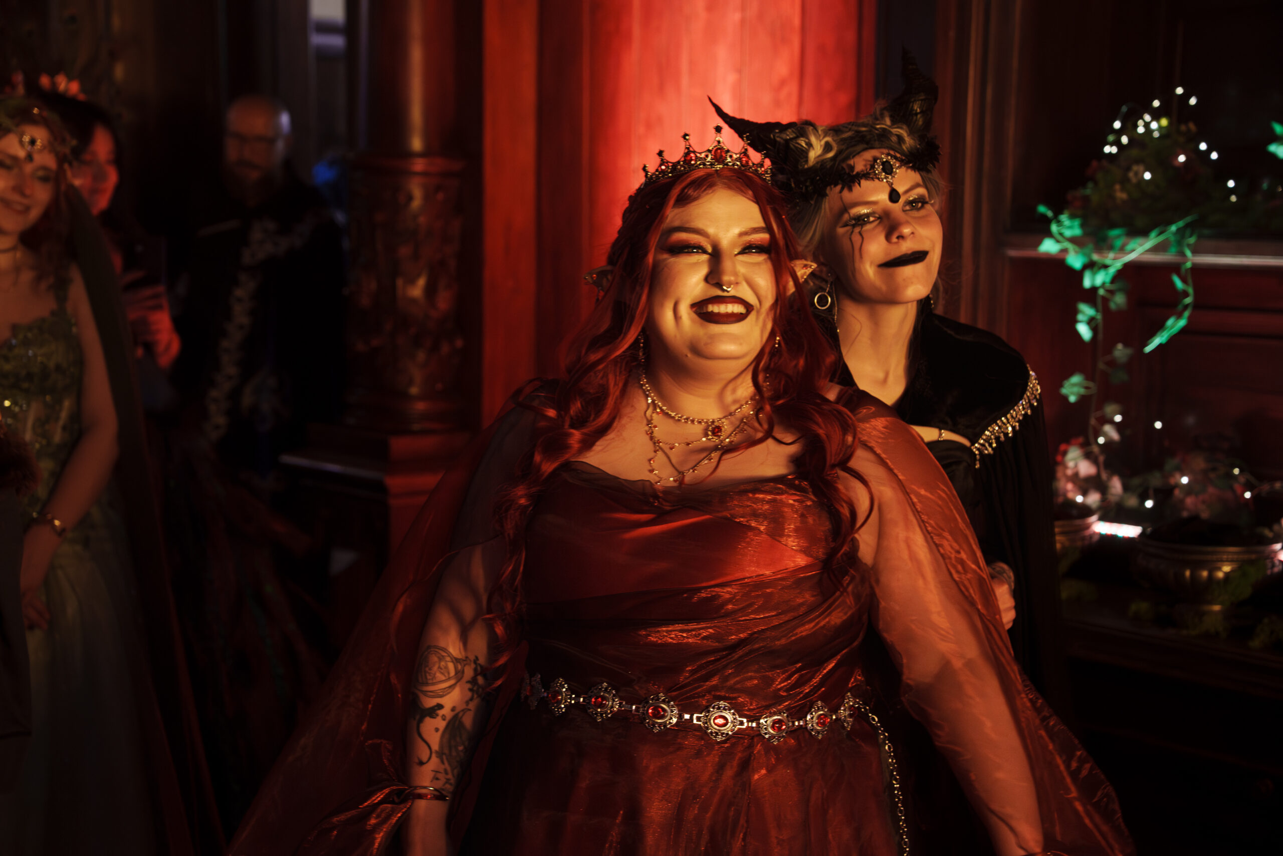 Two women in elaborate costumes pose together, one with a crown and elf ears, wearing a red dress under warm red lighting at a festive event.