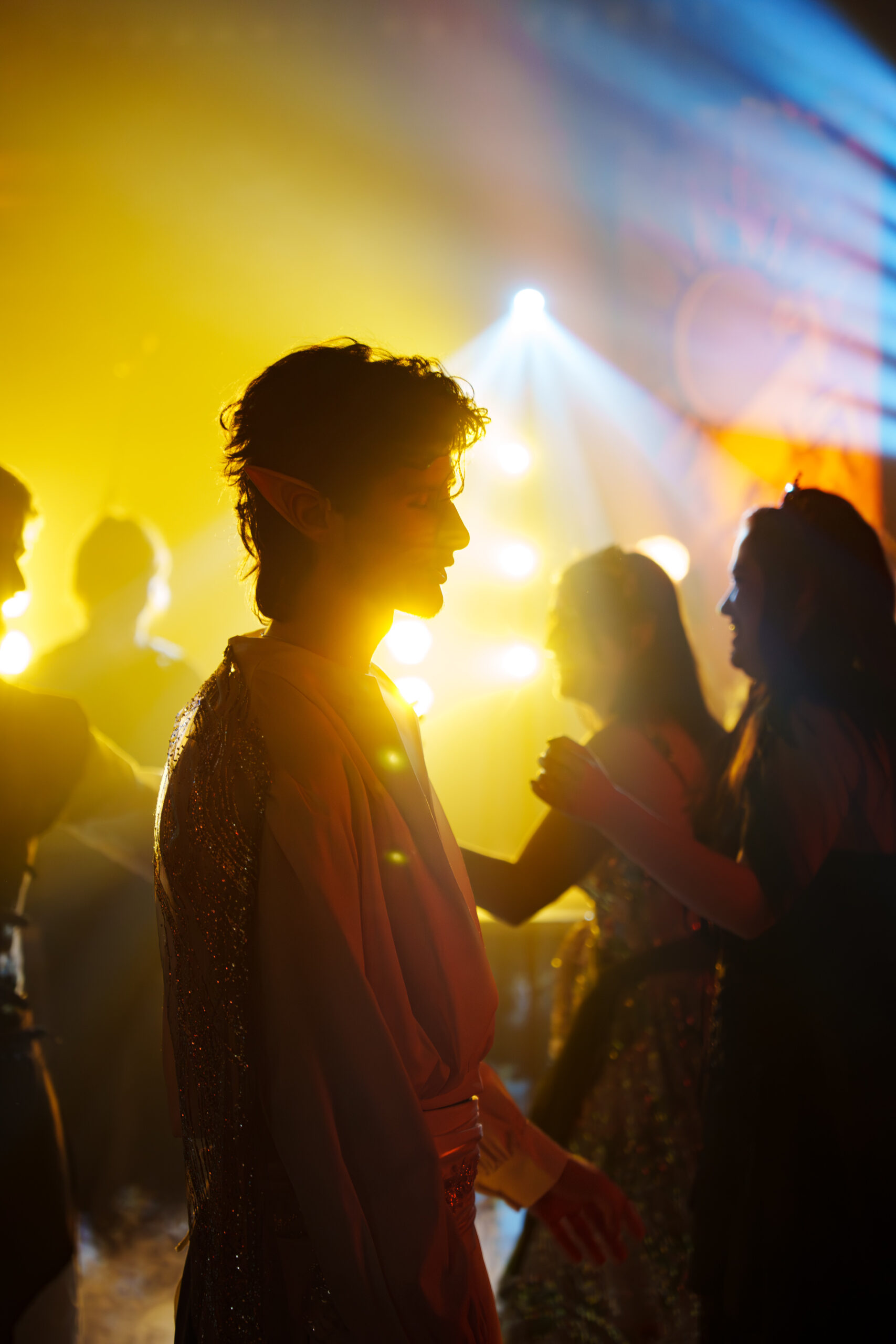 Silhouetted person in a sparkly costume wearing elf ears at a neon-lit party with yellow and blue lights.
