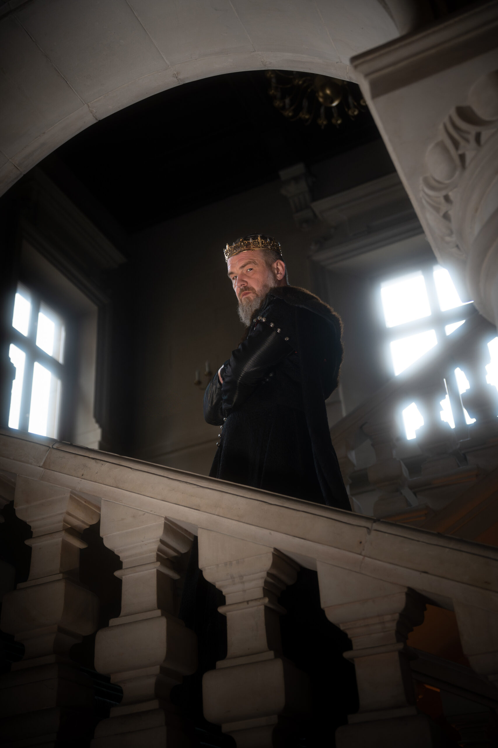 A bearded man wearing a gold crown stands on a marble balcony in a dim, grand interior with arched architecture behind him.