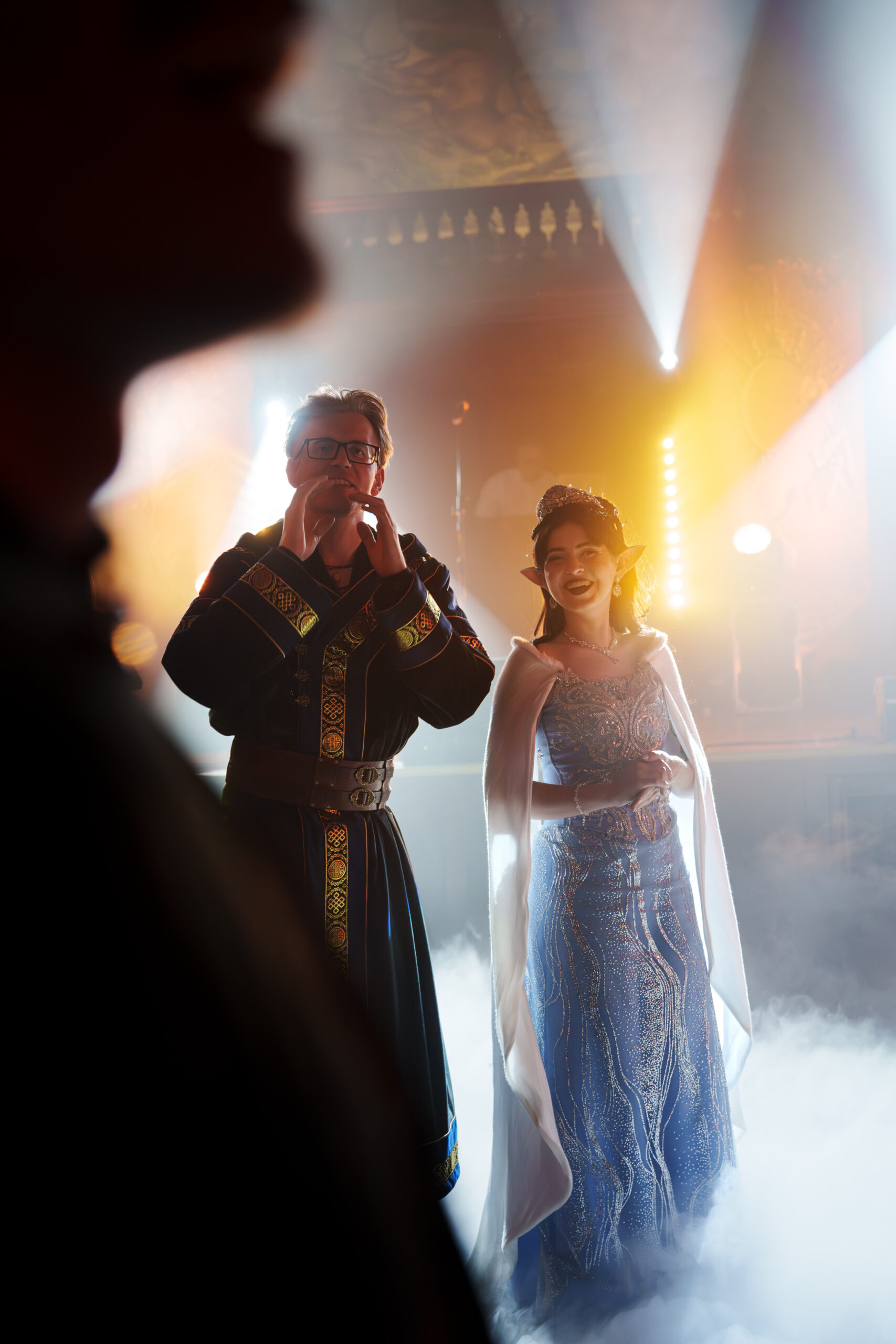 Two costumed performers on a foggy stage, backlit by bright spotlights and warm colors, smiling at the audience.