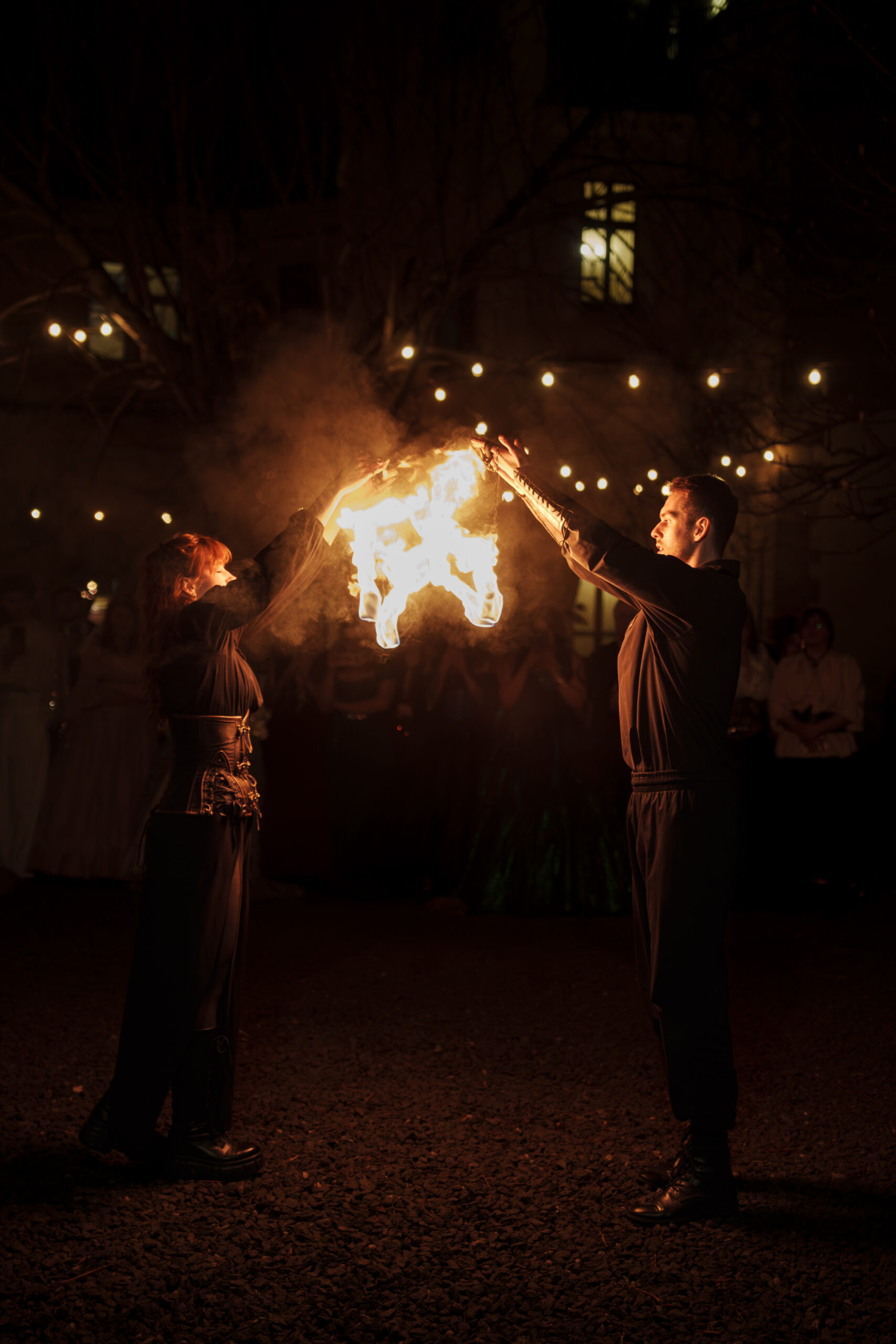 Two performers in dark clothing hold flaming props together, creating a bright fire between them at night under string lights.