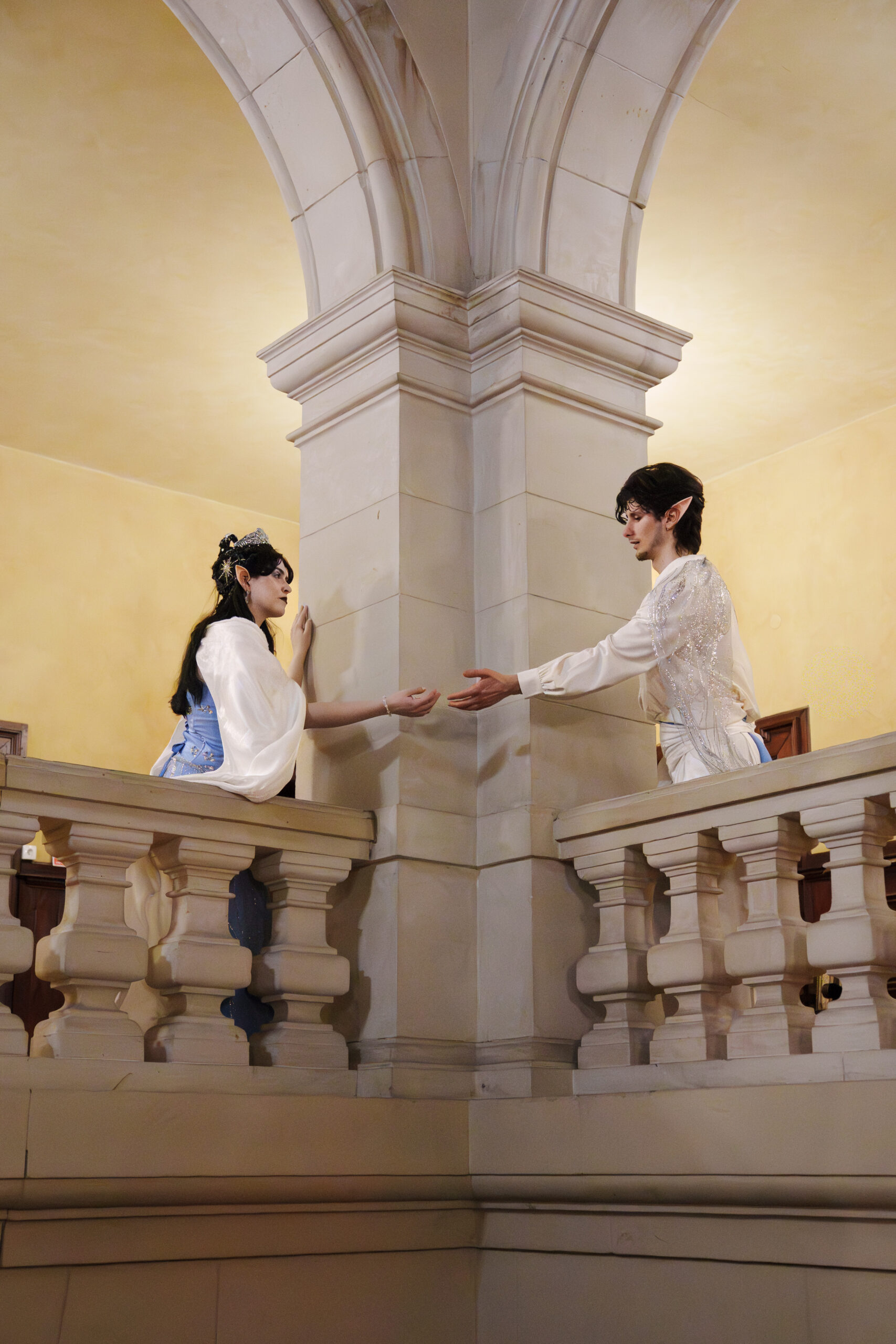Two elf-like performers on a balcony, reaching toward each other, inside a stone-arched hall with beige walls and railing.