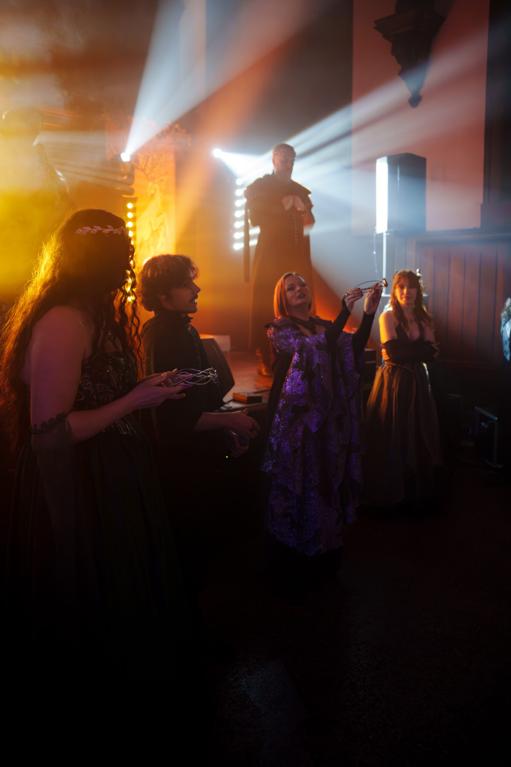 Performer in a dark costume on a stage with bright spotlights, while people in period dresses watch and clap nearby.