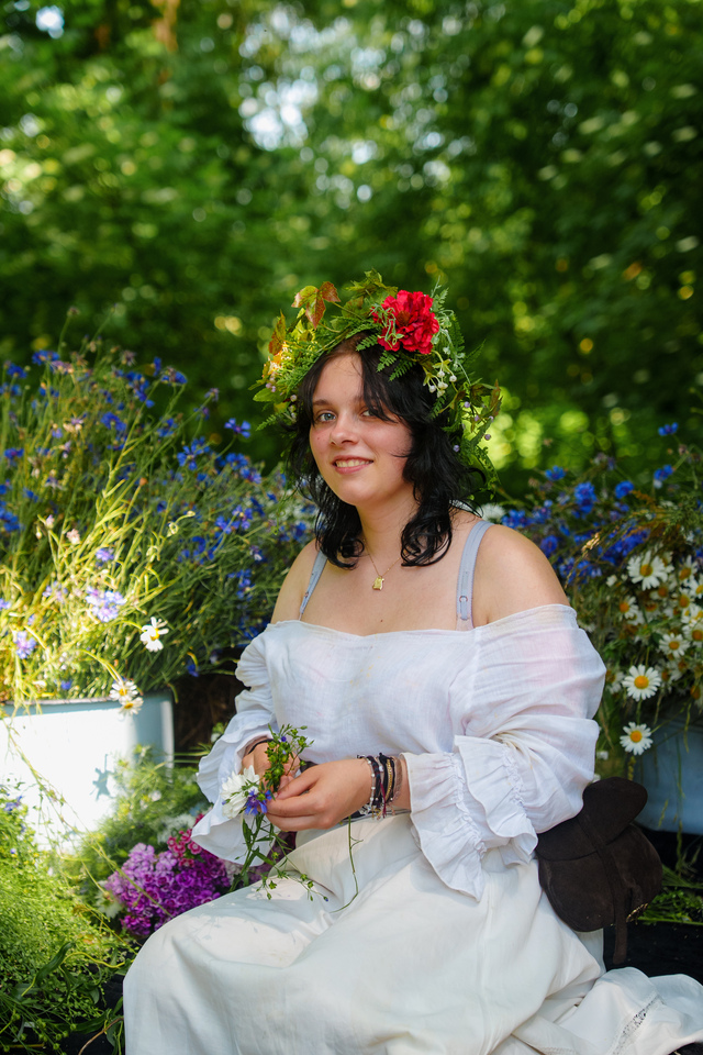 Young woman with a flower crown sitting among blue and white flowers in a garden outside the frame's greenery behind her.