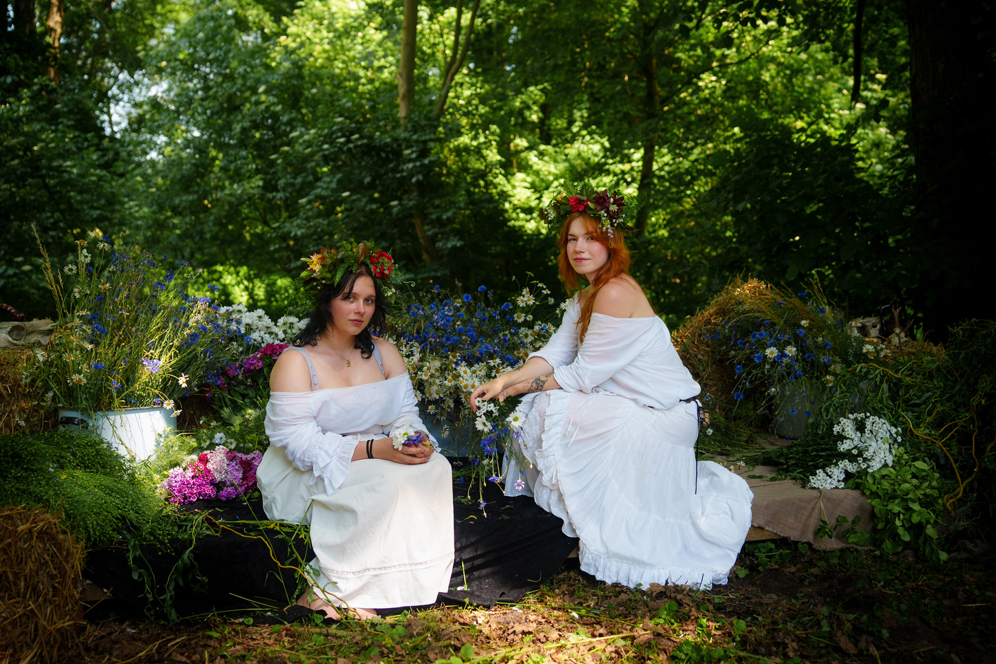 Two women in white dresses with flower crowns sit among a colorful flower display in a sunlit garden park.