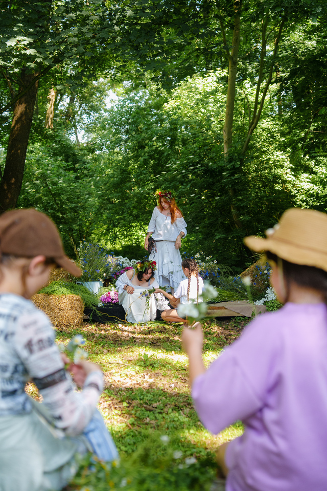 Group of people in a sunlit forest clearing; two hat-wearing participants in the foreground offer flowers to three women in white dresses with flower crowns.