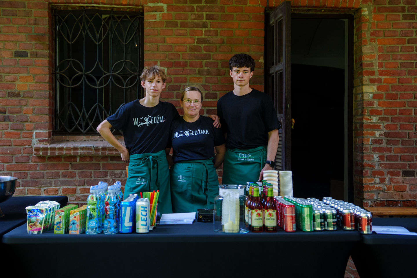 Three volunteers stand behind a long refreshment table with drinks and cups outside a brick building, wearing black shirts and green aprons.