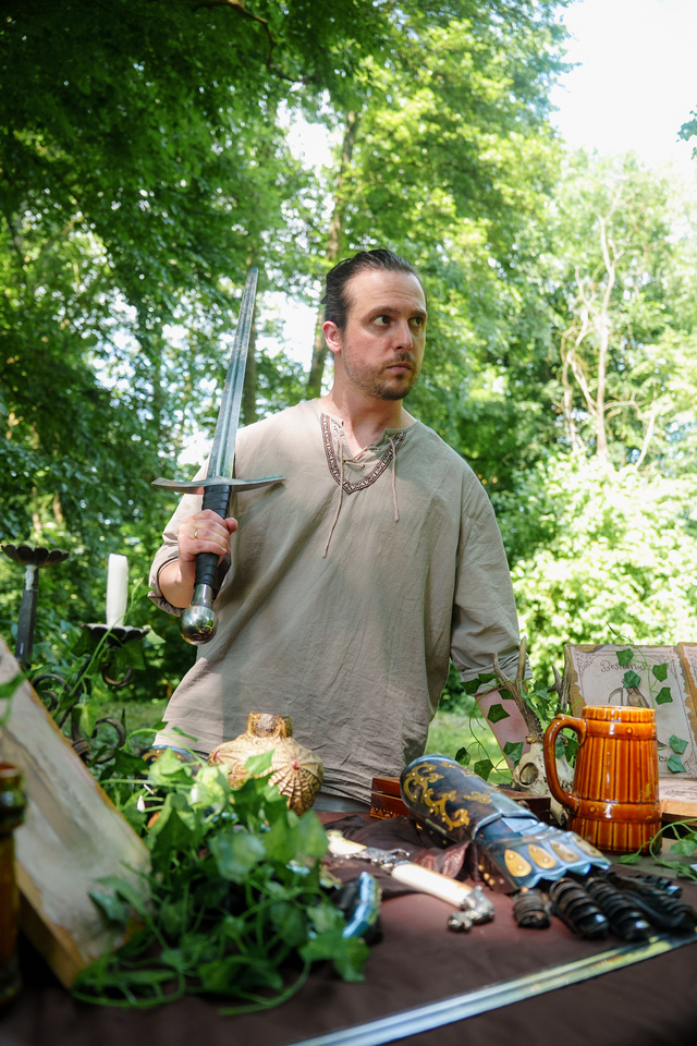 Man in a beige tunic holds a long sword at a table of medieval props during an outdoor fair or market setting.
