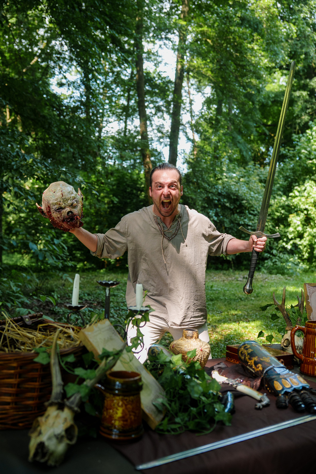 Man in a beige tunic brandishing a sword in one hand and a fake severed head in the other at an outdoor props table in a forest setting.
