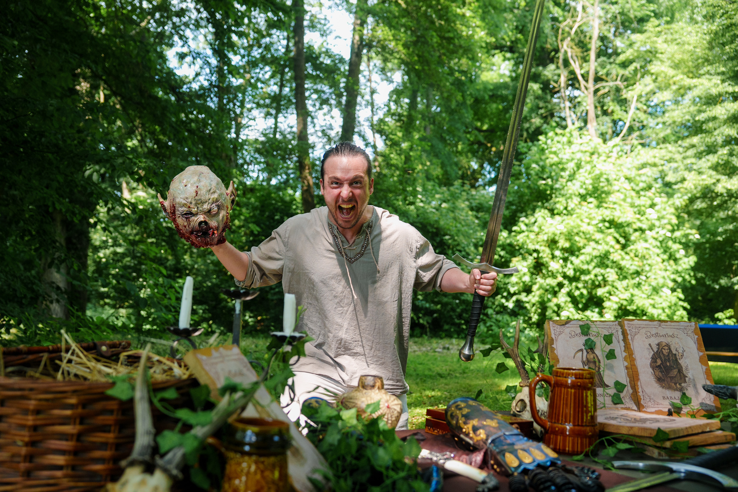 Man in period costume holding a fake decapitated head and a longsword at a medieval-themed outdoor table of props.