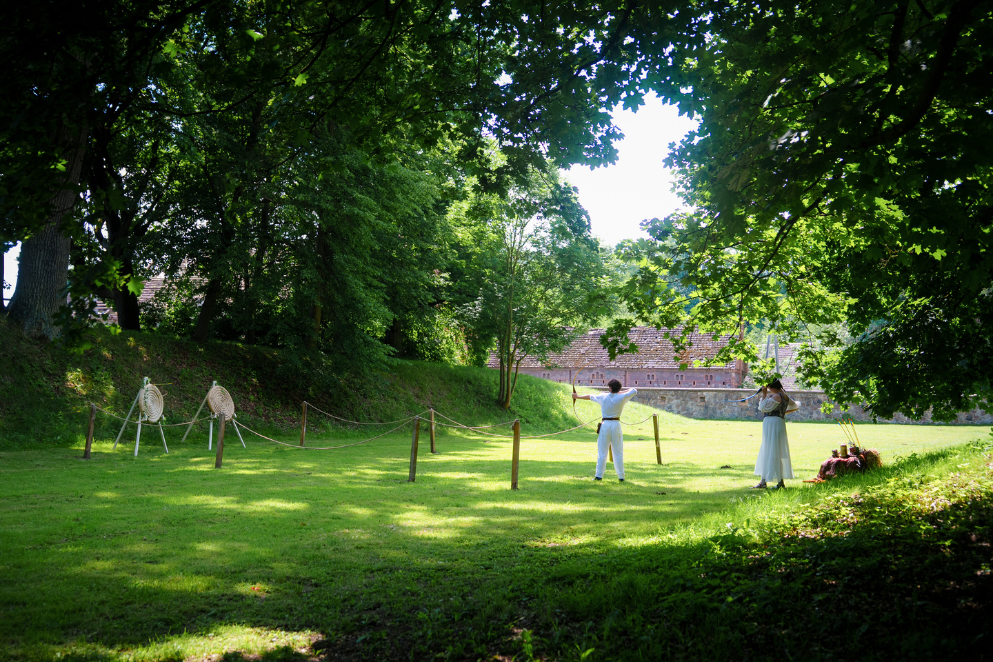 Two people in a sunlit park: a man drawing a bow at left with archery targets nearby and a brick building in the background.