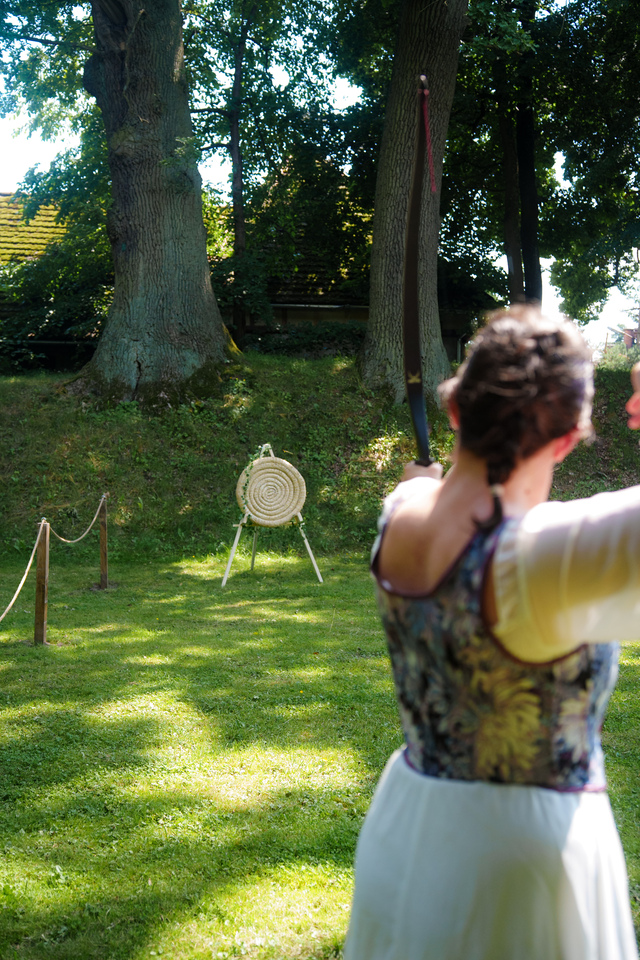 A person outdoors in a park aiming a bow at a round straw archery target on a stand, with trees and sunlight in the background.