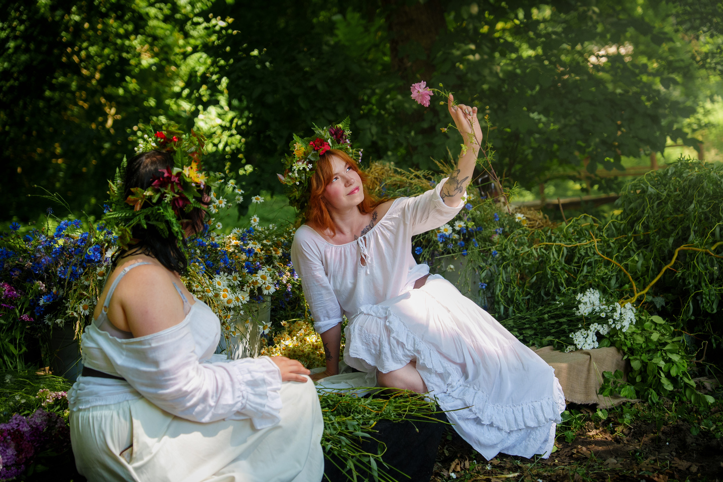 Two women in white dresses with flower crowns sit among blue and white wildflowers in a sunlit garden; one reaches up to pluck a pink bloomVersatile alt text for context: outdoor scene with bohemian attire, flowers, and relaxed activity.