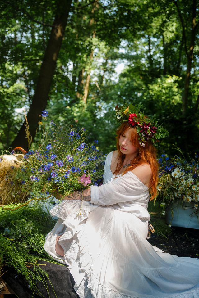 Woman in a flowing white dress and flower crown sits in a sunlit forest, holding a wildflower bouquet.