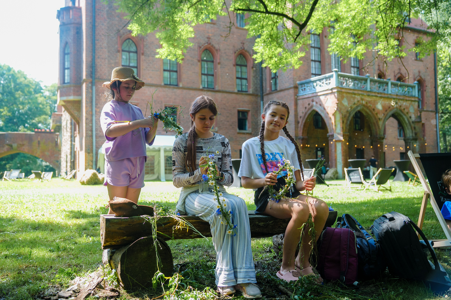 Three girls sit on a wooden bench outdoors, weaving flower garlands with a brick building in the background.