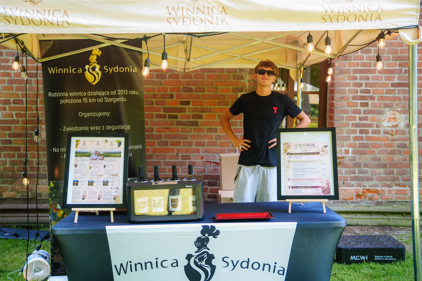 Young man in sunglasses behind a Winnica Sydonia wine stall under a canopy with string lights.