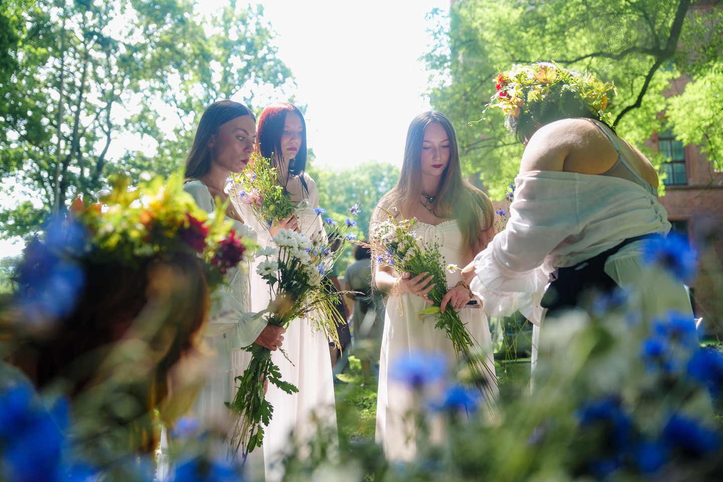 Four women in white dresses standing in a sunlit garden, each holding a bouquet of wildflowers.