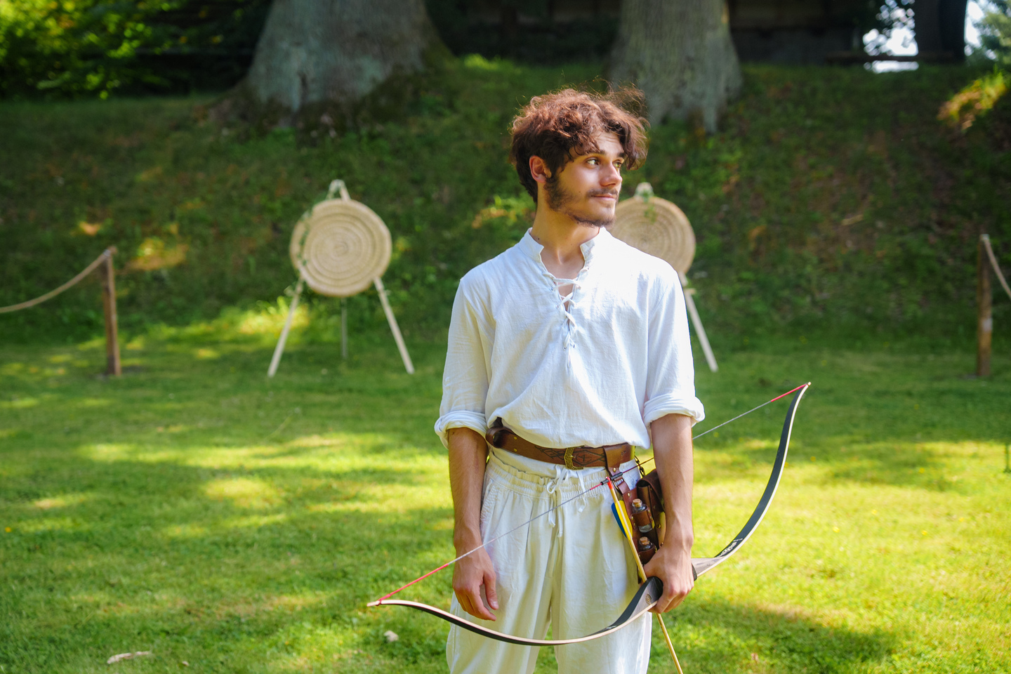 Young man in white medieval-style shirt and belt holds a bow outdoors, with archery targets in the background on a sunny lawn.