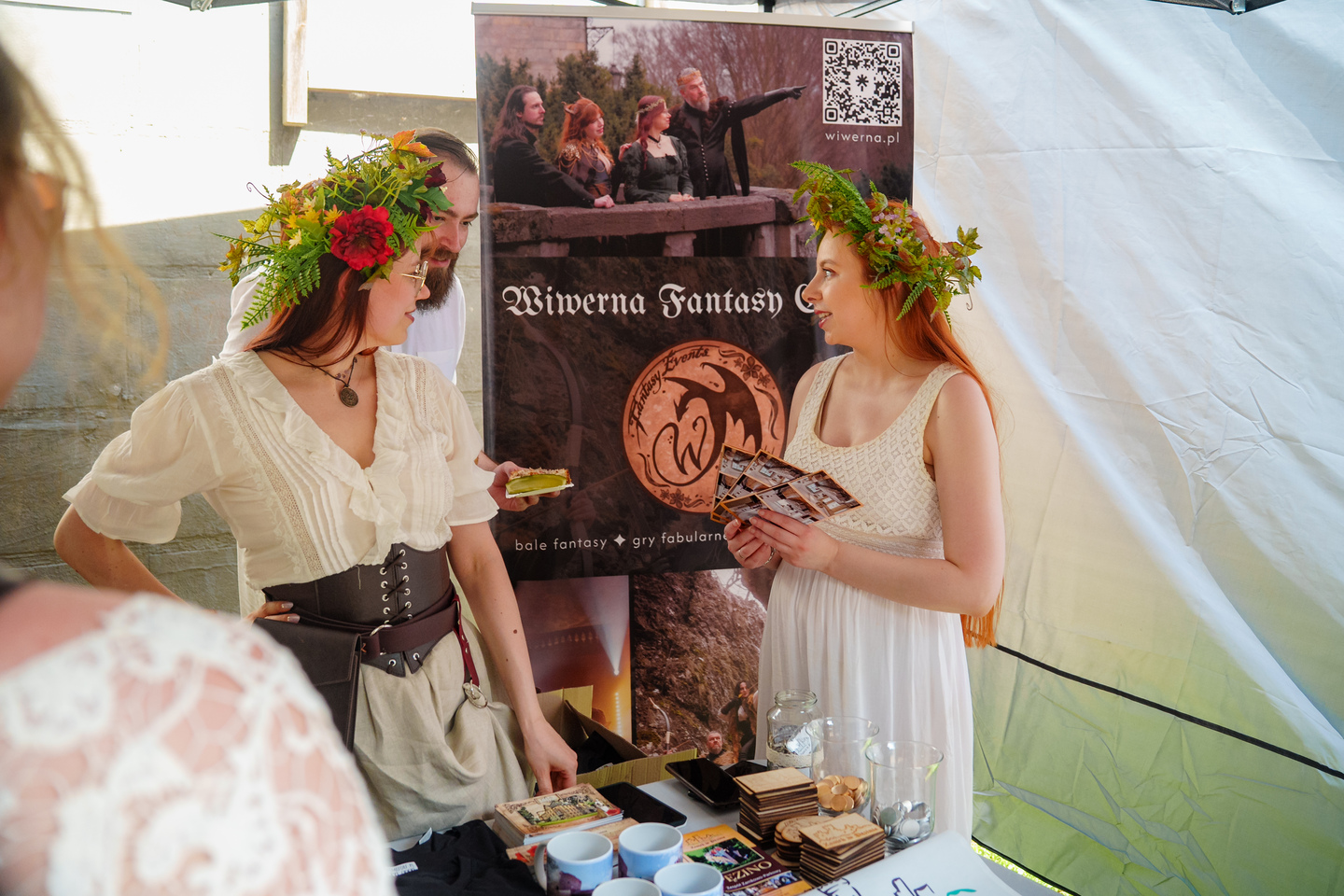 Two women in flower crowns and light dresses chat at a fantasy-themed booth behind a promotional banner.