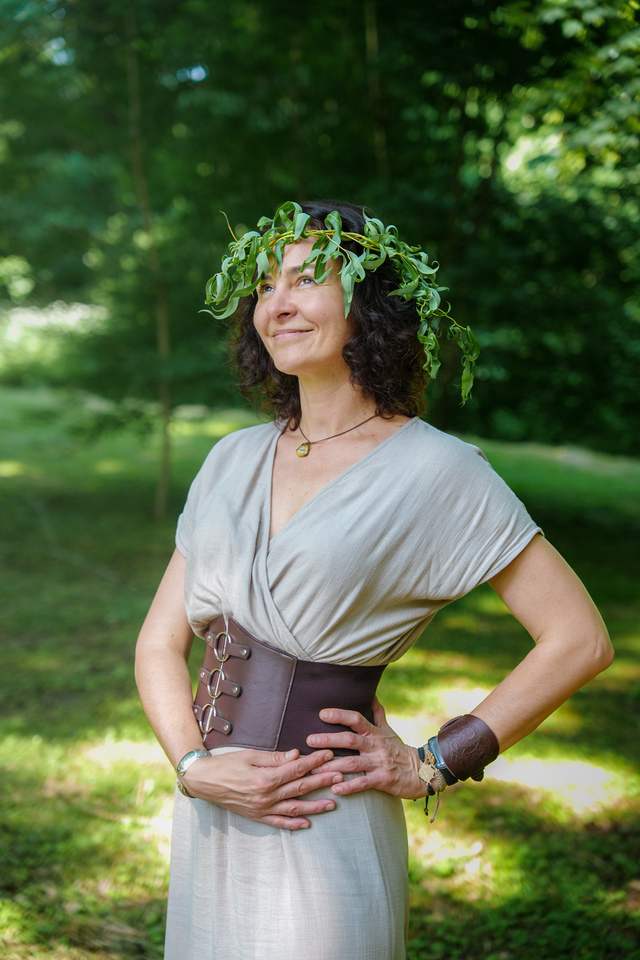 Woman in a light gray dress with a wide brown belt stands outdoors in a forest, wearing a leafy crown and a calm, confident smile.