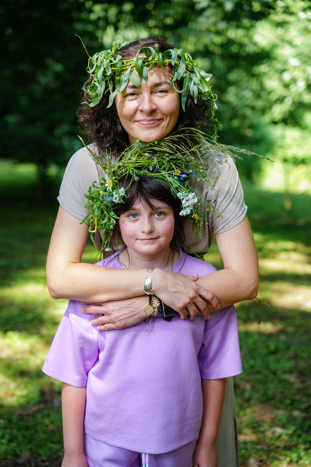 Mother embraces daughter in a park, both wearing leafy flower crowns made of greenery and wildflowers.