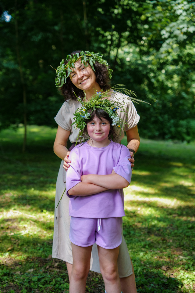 Smiling woman hugs a girl in a park; both wear leafy flower crowns and look toward the camera.
