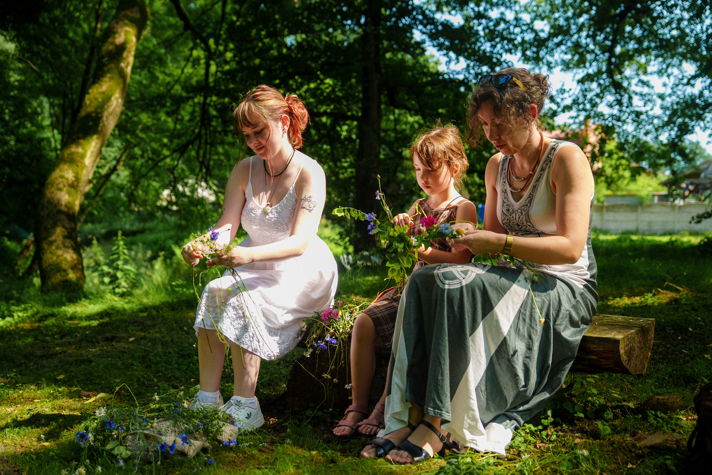 Three people—two adults and a child—sit on a log outdoors, weaving wildflowers into flower garlands in a sunlit park.”] ,