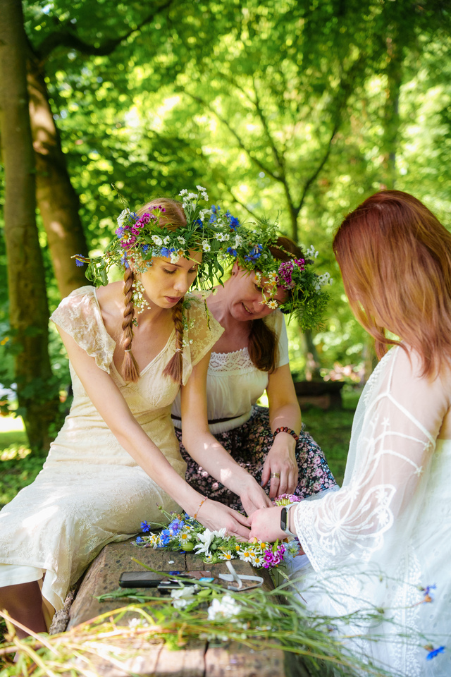 Three women in a sunlit forest weave flower crowns, with one wearing a crown of wildflowers.