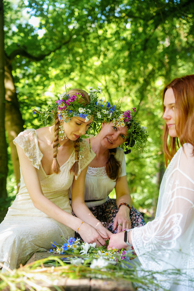 Three women wearing flower crowns craft a floral garland together in a sunlit forest clearing.