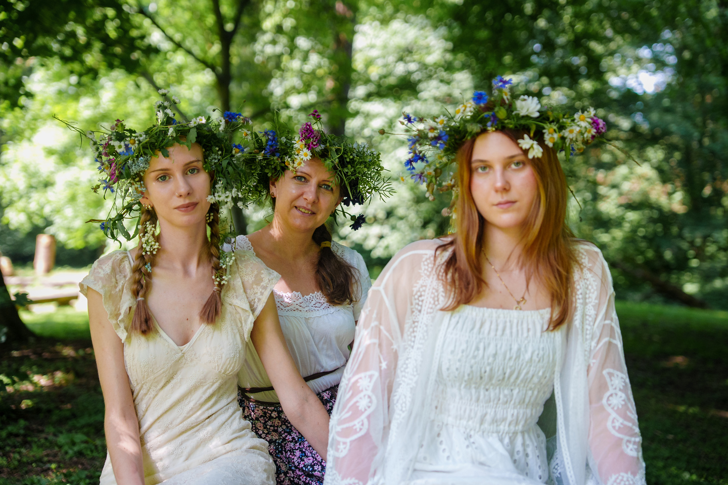 Three women wearing colorful flower crowns sit together in a sunlit park garden.