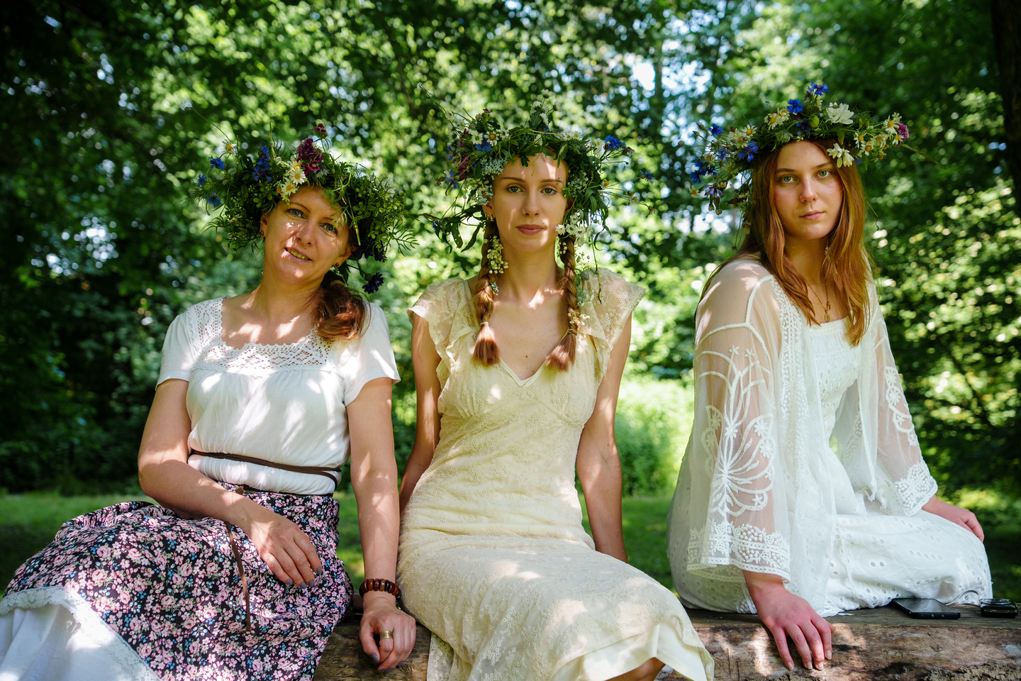 Three women in white dresses with flower crowns sit on a log in a sunlit park.