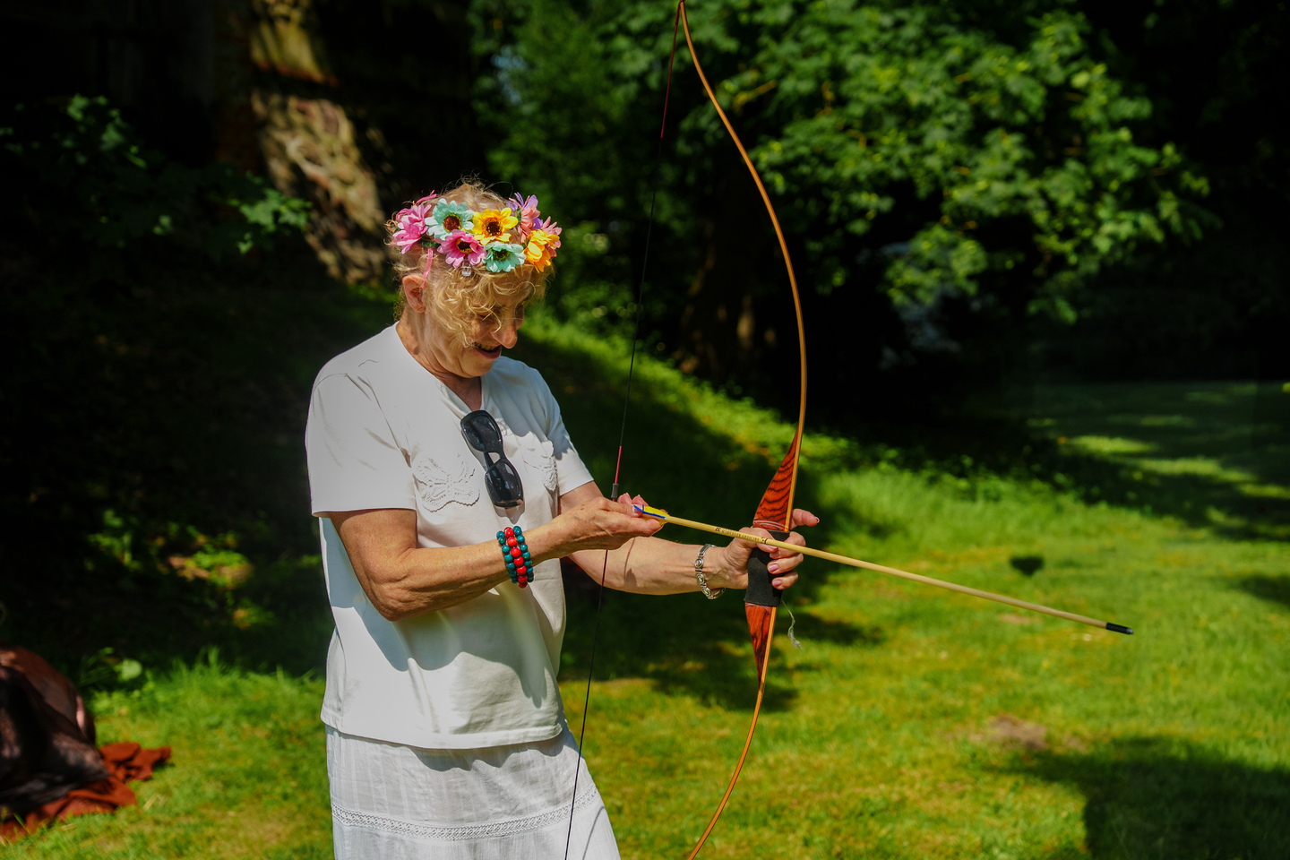 Older woman with a colorful flower crown drawing a bow and aiming an arrow outdoors in a sunny park.