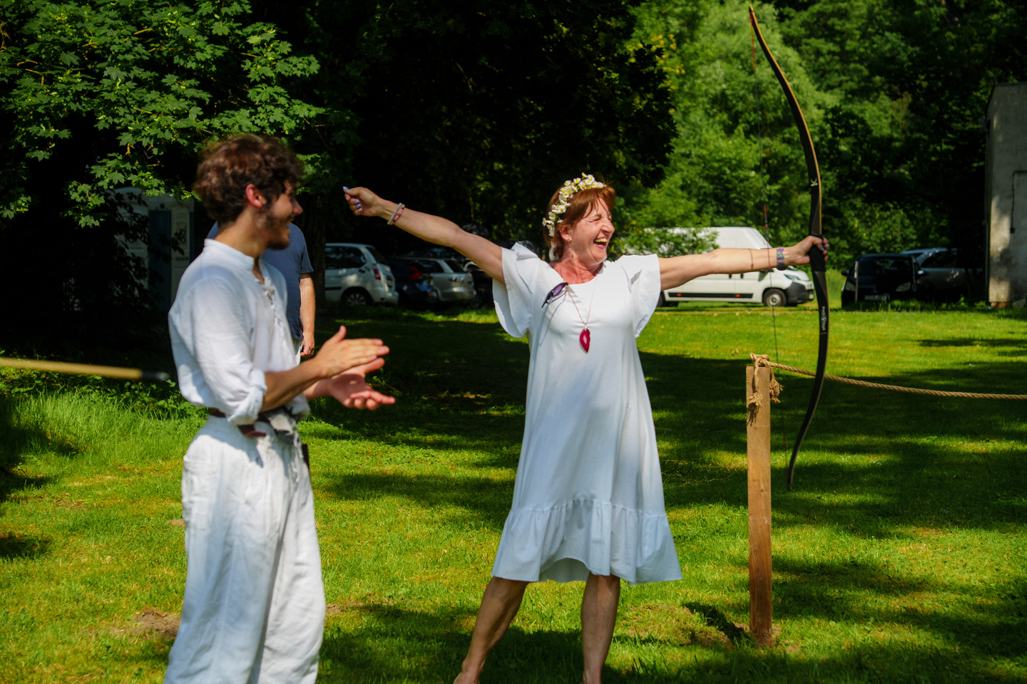 Woman in a white dress with a flower crown draws a bow in a sunny park while a man in white claps beside her.