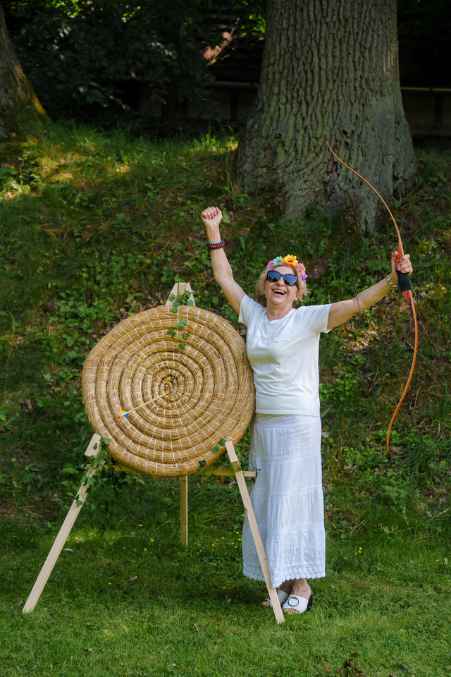 Joyful woman in white with a flower crown and sunglasses raises her fist beside a large woven straw archery target outdoors on grass.