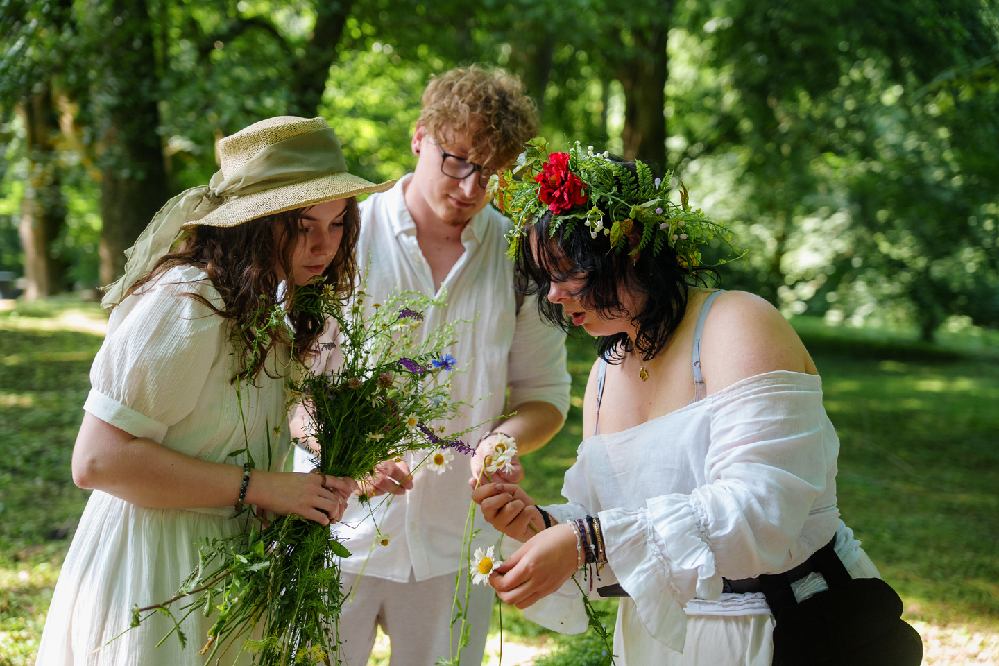 Three friends in a sunlit park wearing white, gathering wildflowers and arranging a flower crown on one person’s head.