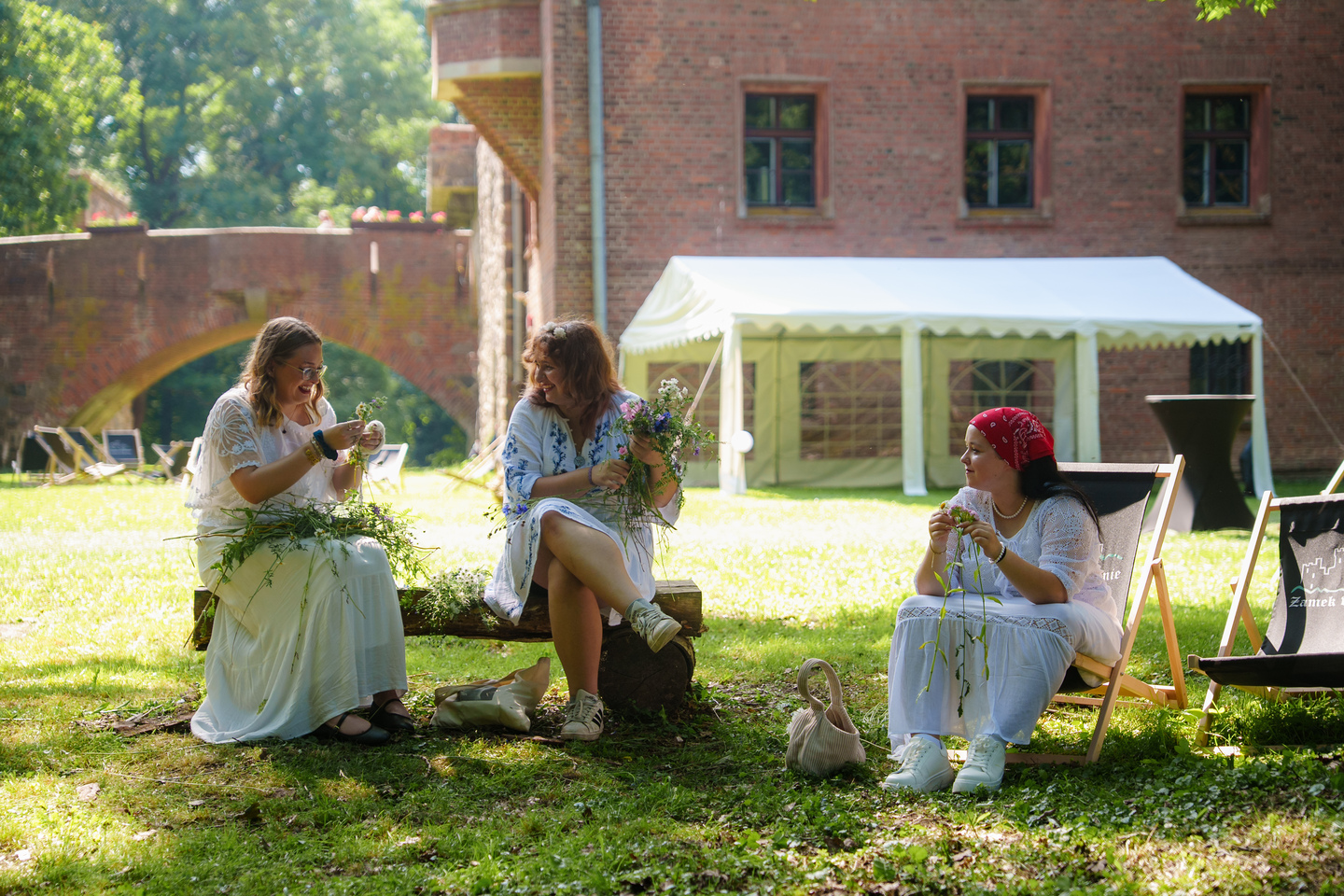 Three women in light dresses sit outdoors on a wooden bench and deck chairs, threading wildflowers into bouquets in a sunny garden near a brick building and white tent.