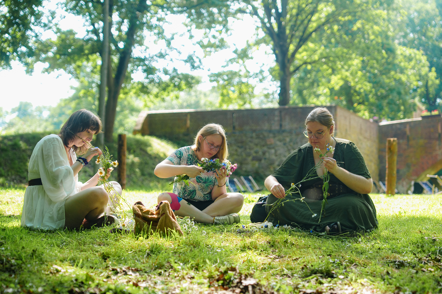 Three women sit on the grass weaving wildflowers into garlands in a sunny park setting.