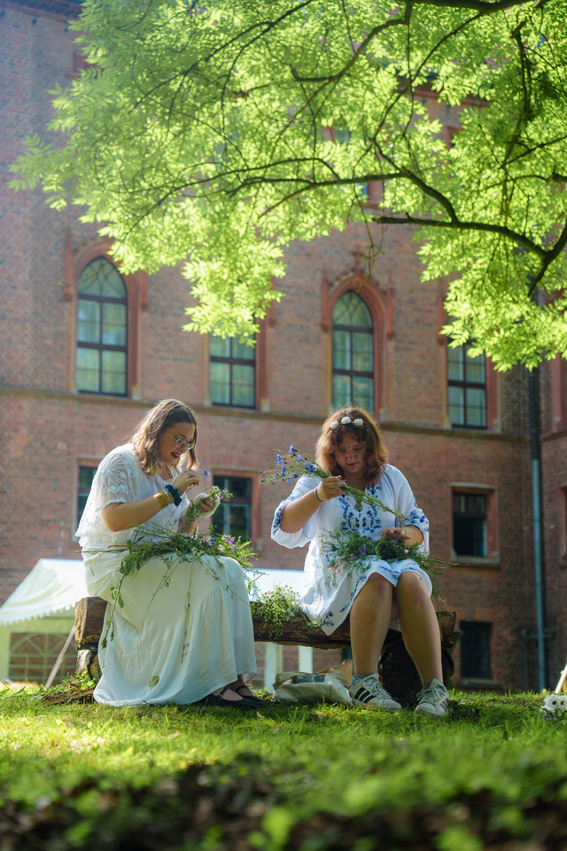 Two women in white dresses sit on a wooden bench weaving wildflowers, outdoors under leafy trees with a brick building in the background.
