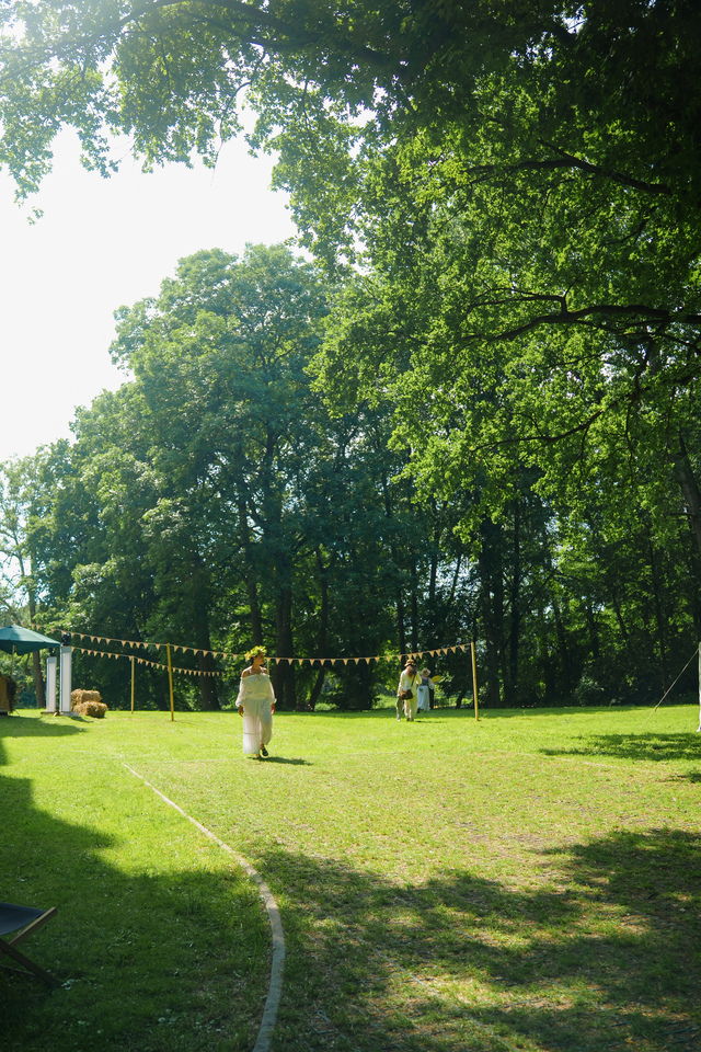 Sunlit grassy park with decorative bunting and a few people walking toward a gathering among tall green trees.