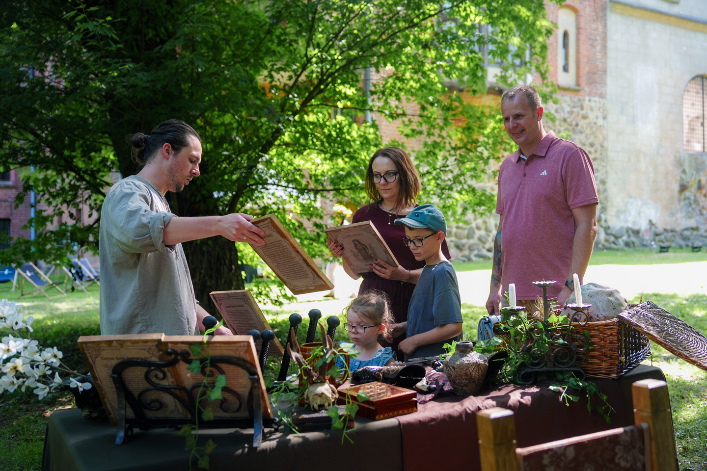 Man showing a vintage book to a family at an outdoor antique stall with various objects on a table under trees.