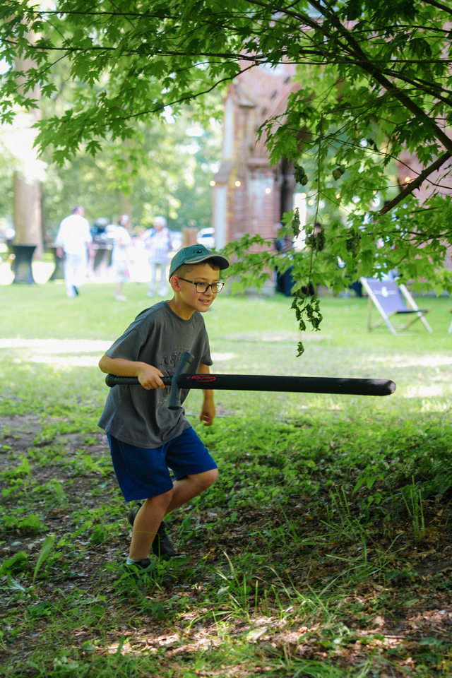 Boy with glasses and a cap runs with a black foam bat in a park under green leaves.