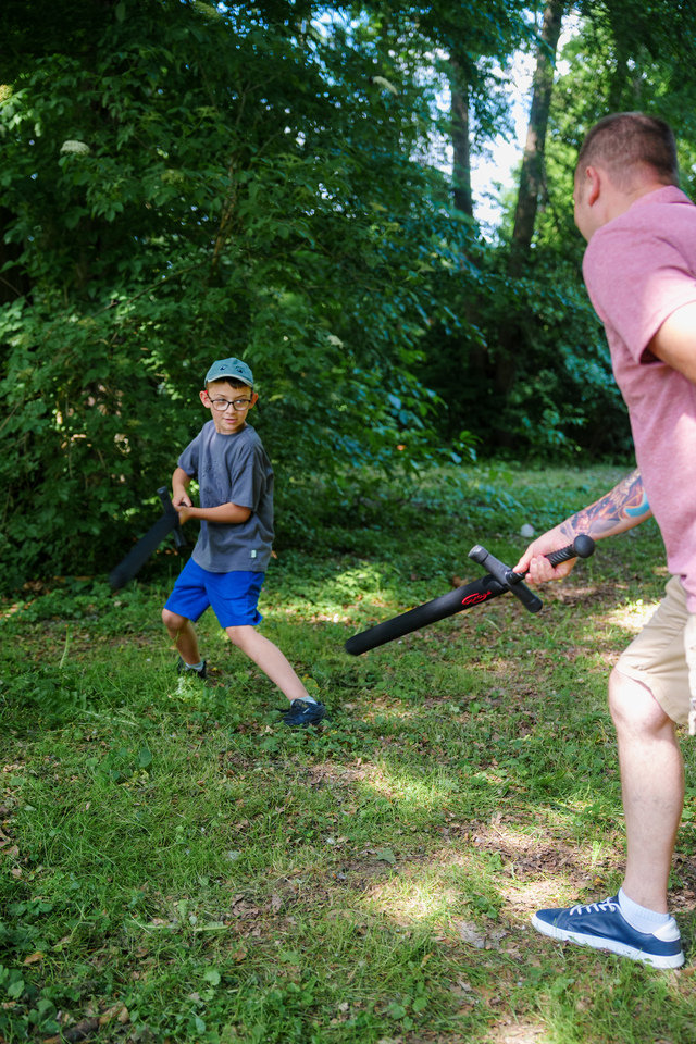 Boy in a gray shirt and blue shorts dodges a foam sword while an adult holds a foam baton in a sunlit park.