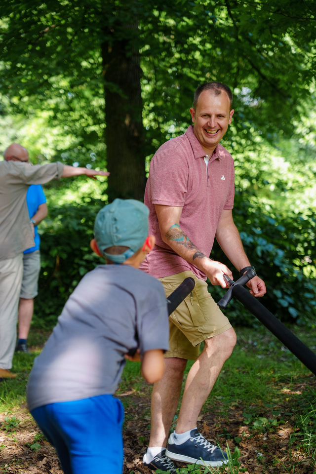 Man in a pink polo shirt smiles while guiding a foam baton in a park as a child approaches; others are in the background among trees and greenery.