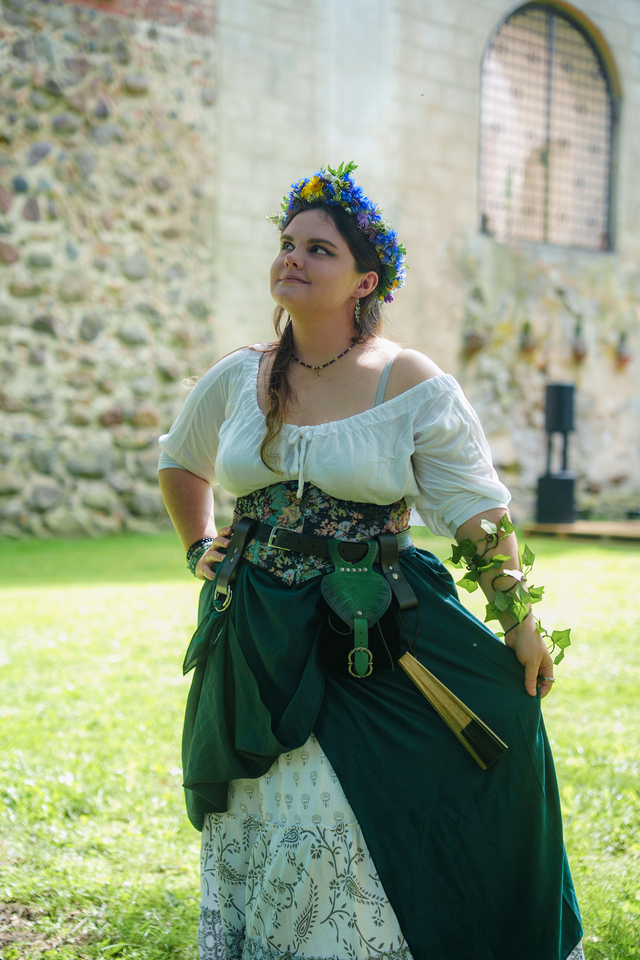 Woman in a Renaissance-style dress with a floral belt and green skirt, wearing a blue-yellow flower crown, outdoors by a stone wall.