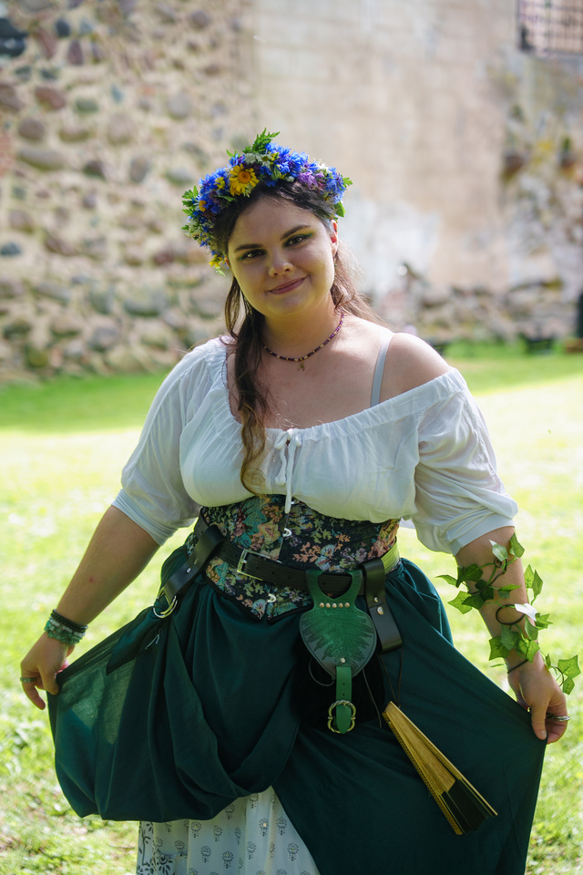 Woman in a floral crown and medieval-style outfit outdoors near an old stone wall, posing for the camera.