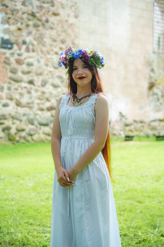 Young woman in a white dress with a colorful flower crown standing on green grass with a stone wall behind her.