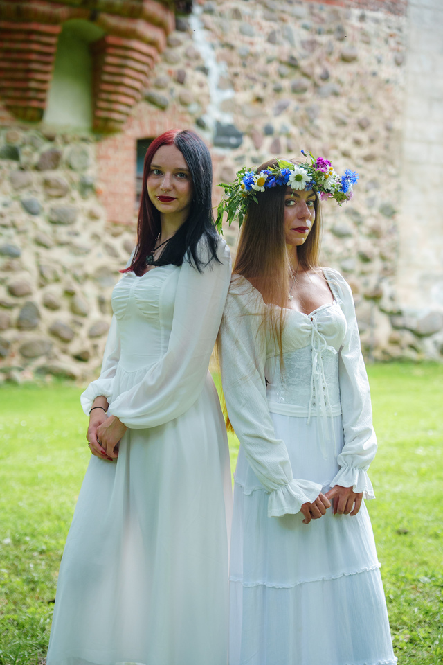Two women in white dresses pose outdoors by a stone wall; one wears a flower crown and bright lipstick.