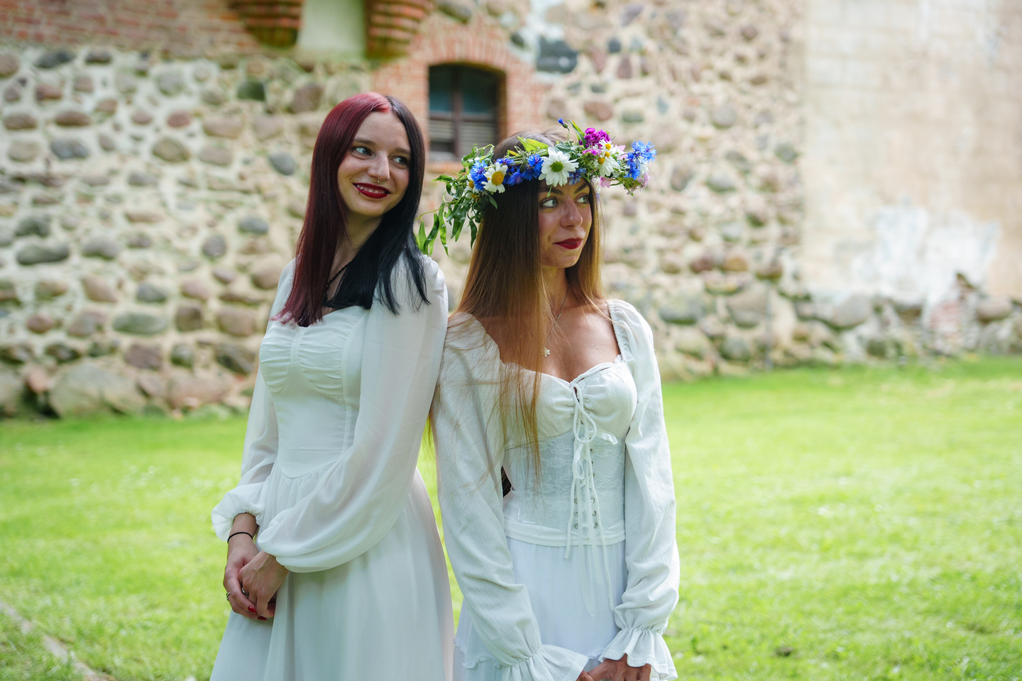 Two women in white dresses pose outdoors by a stone wall, one wearing a colorful flower crown and both smiling.