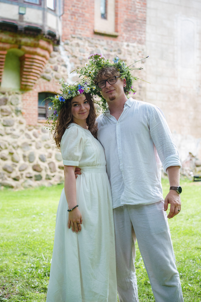 Couple wearing white outfits with flower crowns, smiling and embracing on a grassy lawn outdoors.